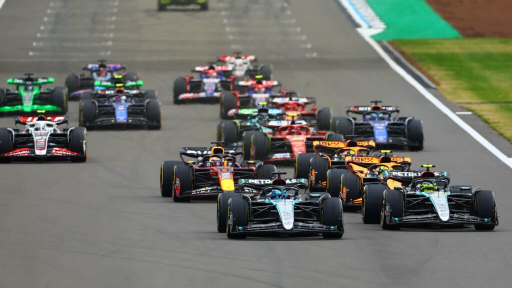 Formula 1 cars racing through Silverstone Circuit during the British Grand Prix, with fans watching from the stands and streaming devices.